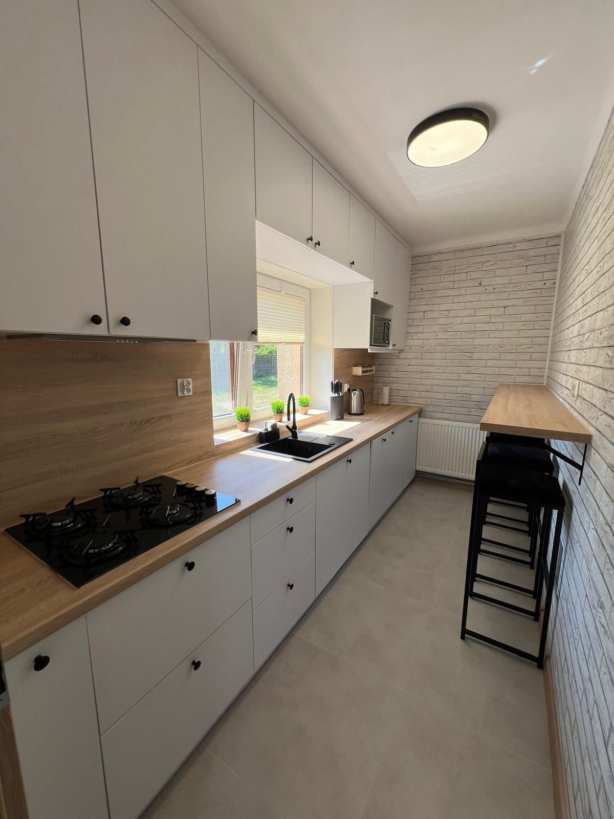 Jasna, nowoczesna kuchnia z białymi szafkami, drewnianym blatem i ceglaną ścianą. Widoczny zlew, płyta gazowa i wysoki stół z krzesłami barowymi.