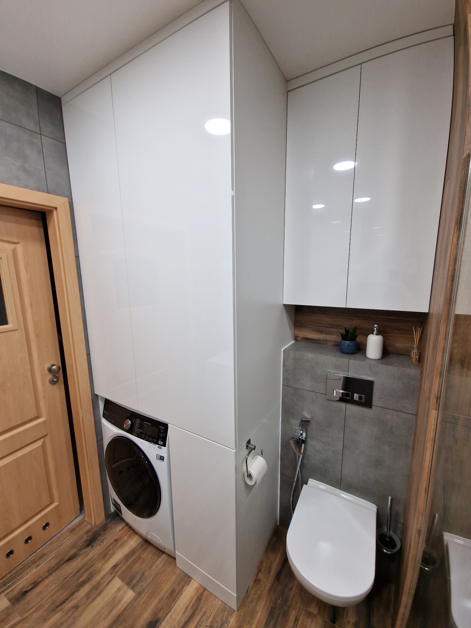 Biała zabudowa meblowa w łazience z pralką, szafkami wiszącymi i toaletą, na tle drewnopodobnych płytek podłogowych i szarej ściany.