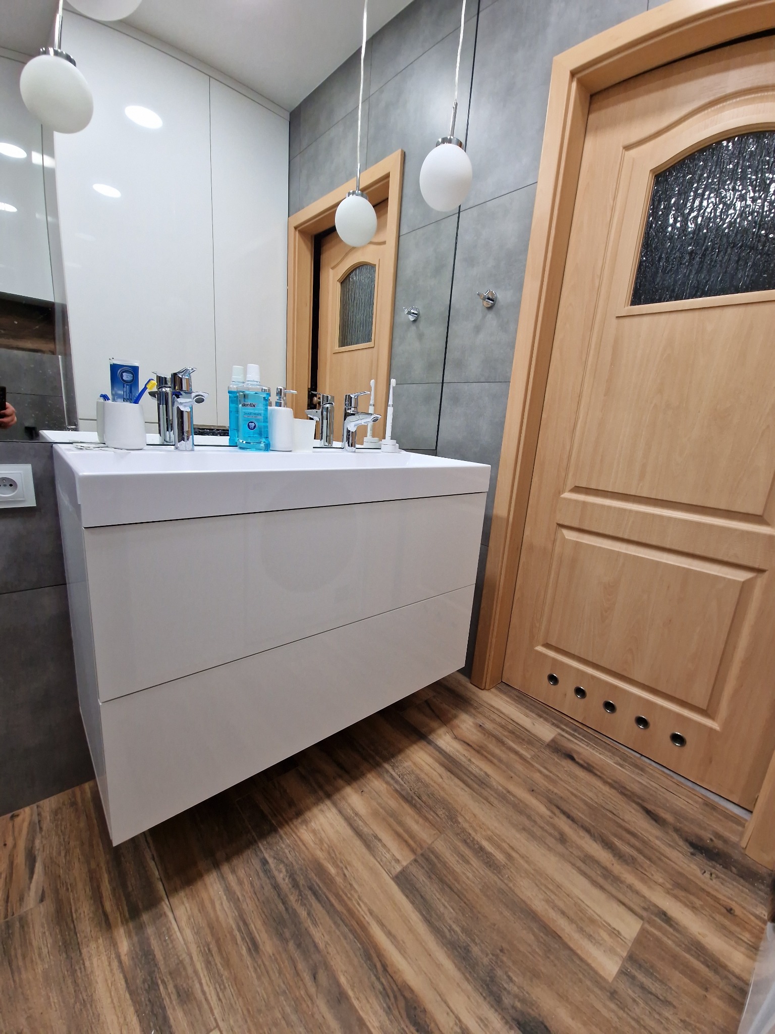 Nowoczesna łazienka z białą szafką pod umywalkę, drewnianą podłogą i drzwiami. Widoczne oświetlenie punktowe i szara ściana z płytkami. Elementy armatury.