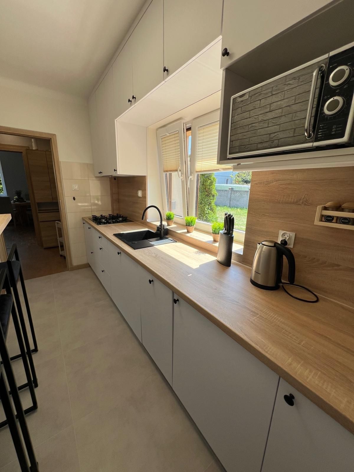 Jasna kuchnia z drewnianym blatem i białymi szafkami, czarny zlew i bateria. Na blacie czajnik i akcesoria kuchenne. Mikrofala nad oknem.