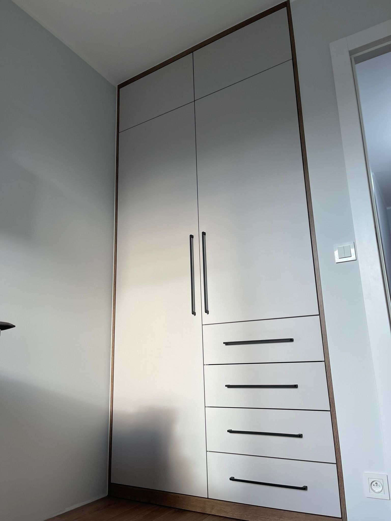 Wysoka, zabudowana szafa wnękowa z szarymi frontami, ciemnymi uchwytami i drewnianym obramowaniem. Widoczne cztery szuflady w dolnej części i dwudrzwiowa szafa powyżej.