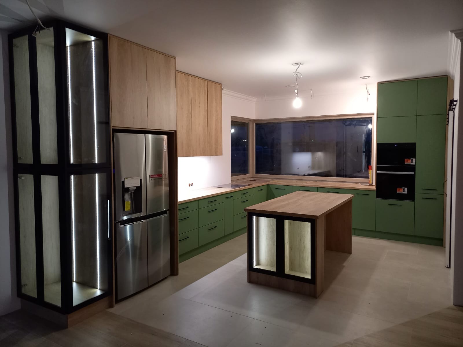 Nowoczesna kuchnia z zielonymi szafkami i wyspą z podświetleniem LED. Duże okno nad blatem roboczym. Obok lodówka side-by-side i przeszklona witryna z oświetleniem.
