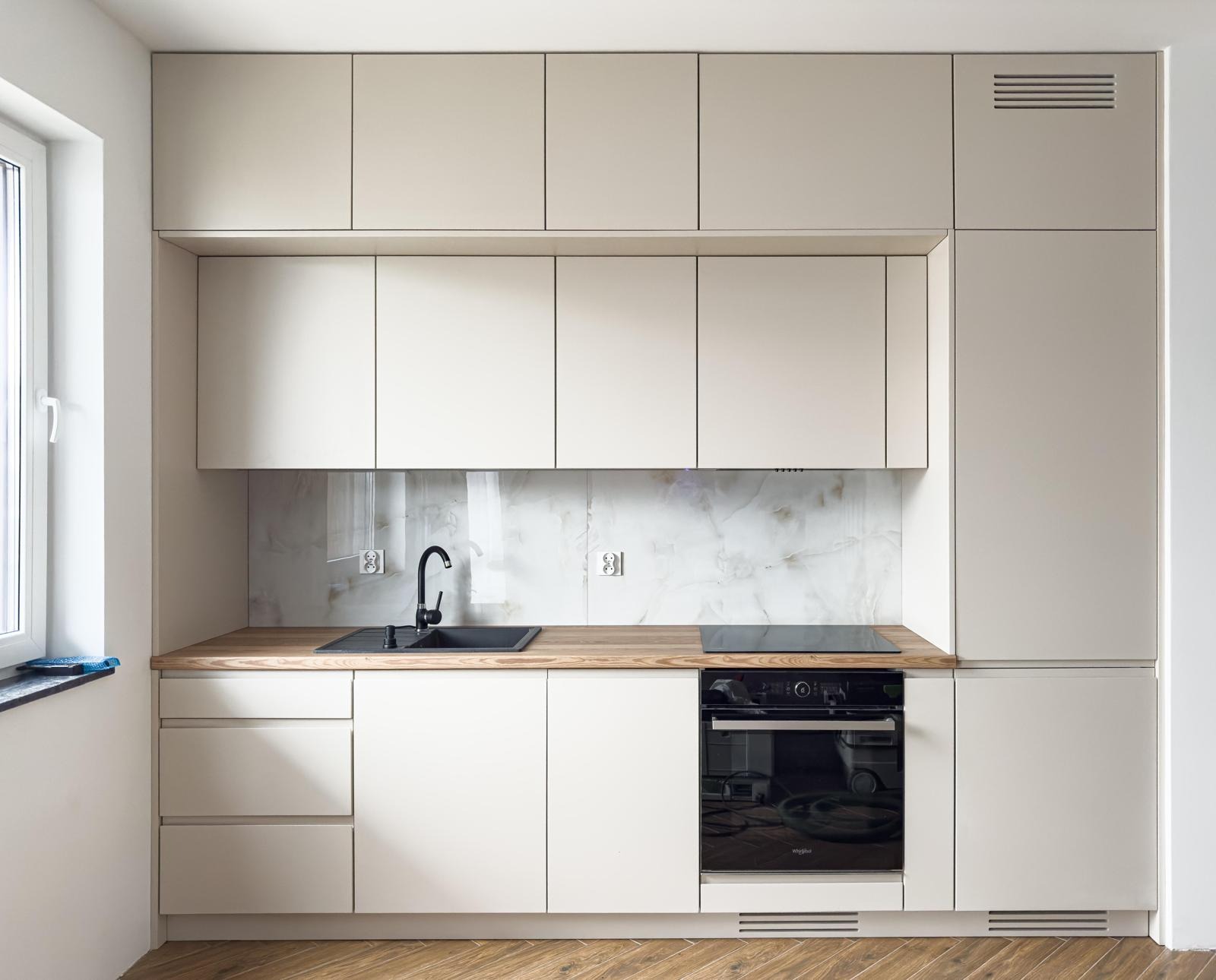 Jasna kuchnia z drewnianym blatem i marmurowym panelem. Górne szafki sięgają sufitu, a czarny zlew i płyta grzewcza kontrastują z jasnym tłem.