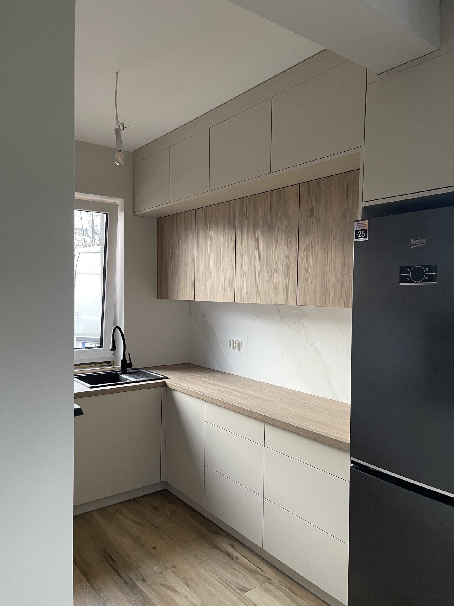 Jasna kuchnia z drewnianymi akcentami: beżowe szafki górne i dolne, drewniany blat, marmurowe wykończenie ściany, czarna bateria i lodówka Beko z naklejką -25.