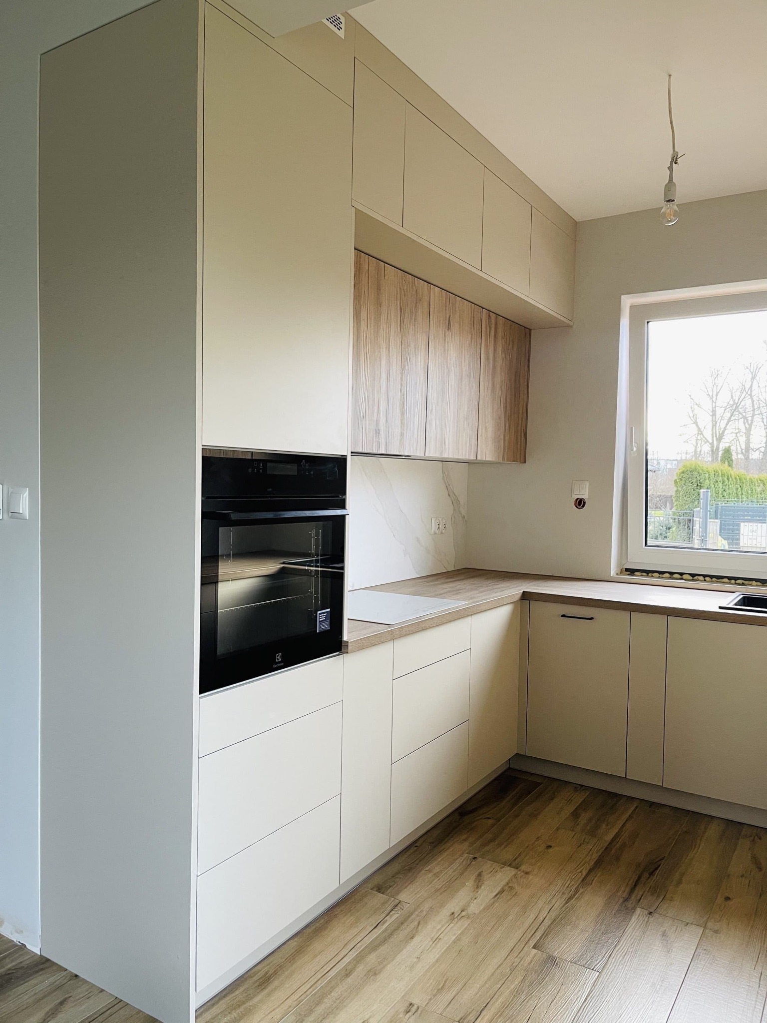 Jasna kuchnia z drewnianymi akcentami: zabudowa z piekarnikiem, szafki wiszące i blat. Parkiet na podłodze, okno z widokiem na zieleń. Minimalistyczny design.