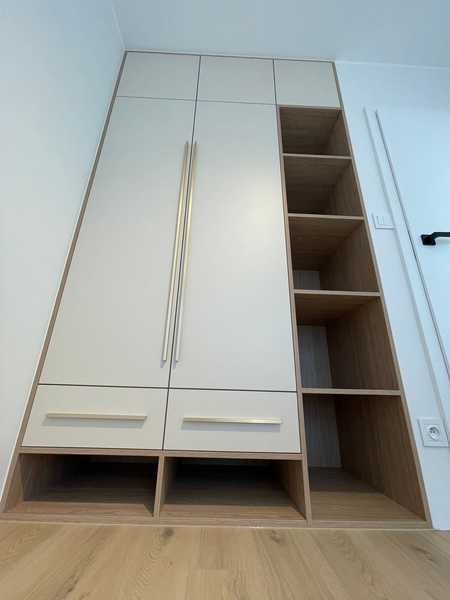 Biała szafa wnękowa na wymiar z drewnianymi półkami i złotymi uchwytami, dopasowana do skosu ściany, widok z dołu, na drewnianej podłodze.
