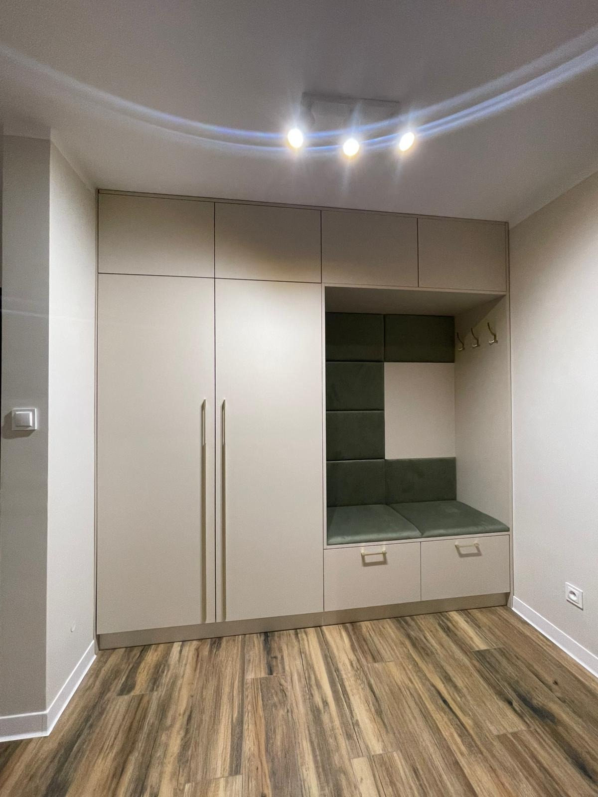 Jasna szafa wnękowa z siedziskiem i wieszakami, wykończona panelami w odcieniach zieleni. Podłoga z paneli imitujących drewno. Oświetlenie sufitowe.