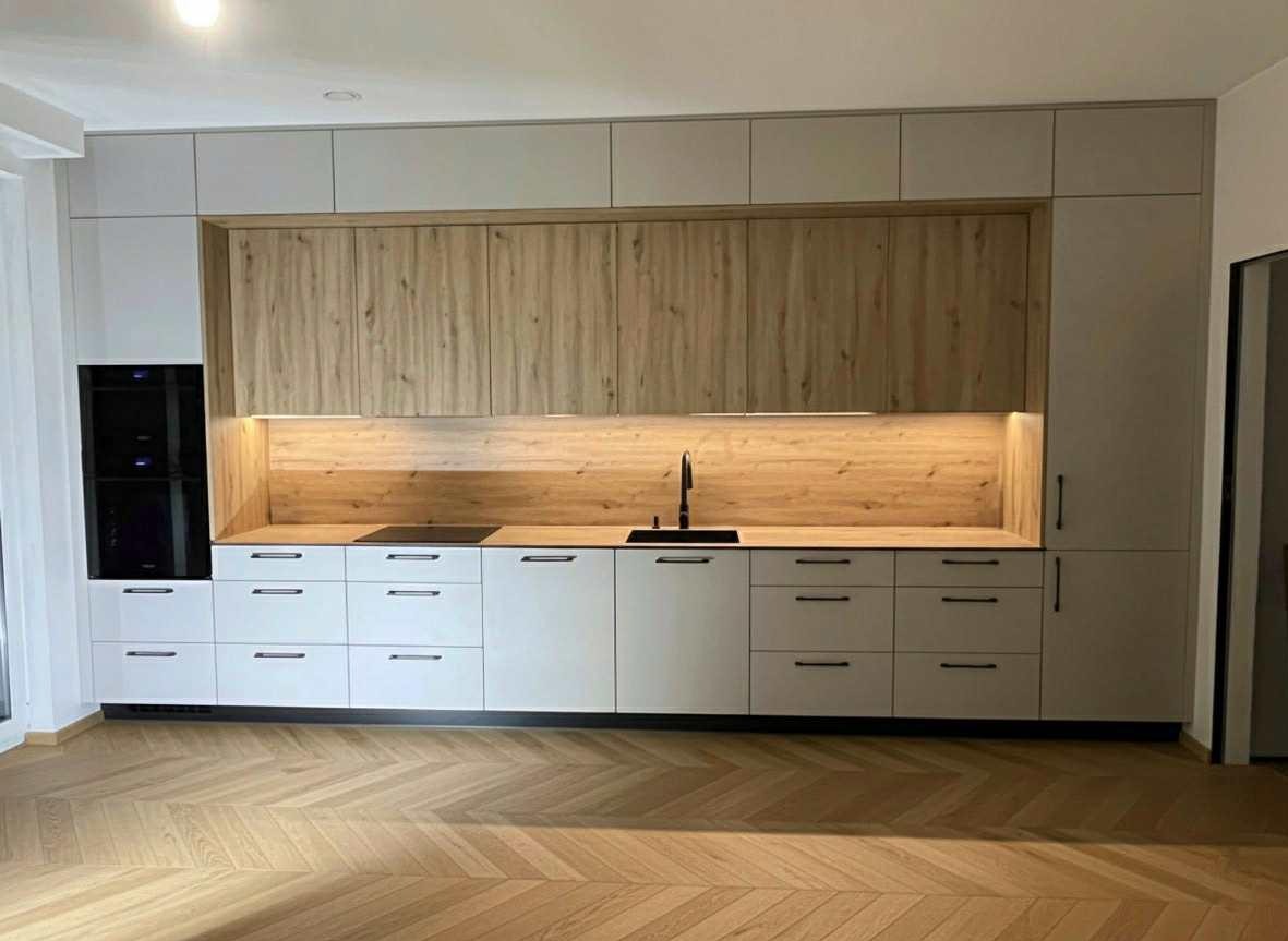 Nowoczesna kuchnia z drewnianymi frontami szafek górnych i białymi dolnymi, oświetleniem LED pod szafkami i parkietem w jodełkę na podłodze.