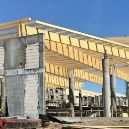 Marcin Maliński - Budowa budynku aquaparku w stanie surowym otwartym: betonowe ściany, drewniana konstrukcja dachu, pracownicy budowlani i żuraw na tle błękitnego nieba.