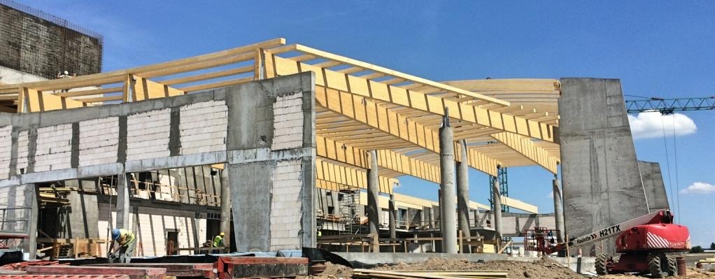 Budowa budynku aquaparku w stanie surowym otwartym: betonowe ściany, drewniana konstrukcja dachu, pracownicy budowlani i żuraw na tle błękitnego nieba.