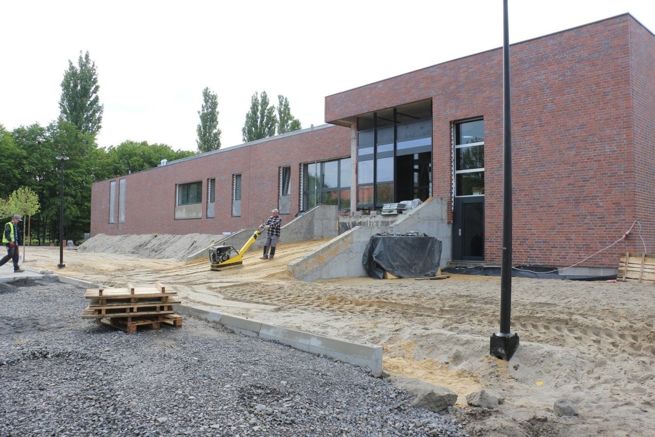 Budowa aquaparku: murowany budynek z dużymi oknami, pracownik zagęszcza piasek zagęszczarką. Palety i gruz leżą na ziemi. Widok na plac budowy.