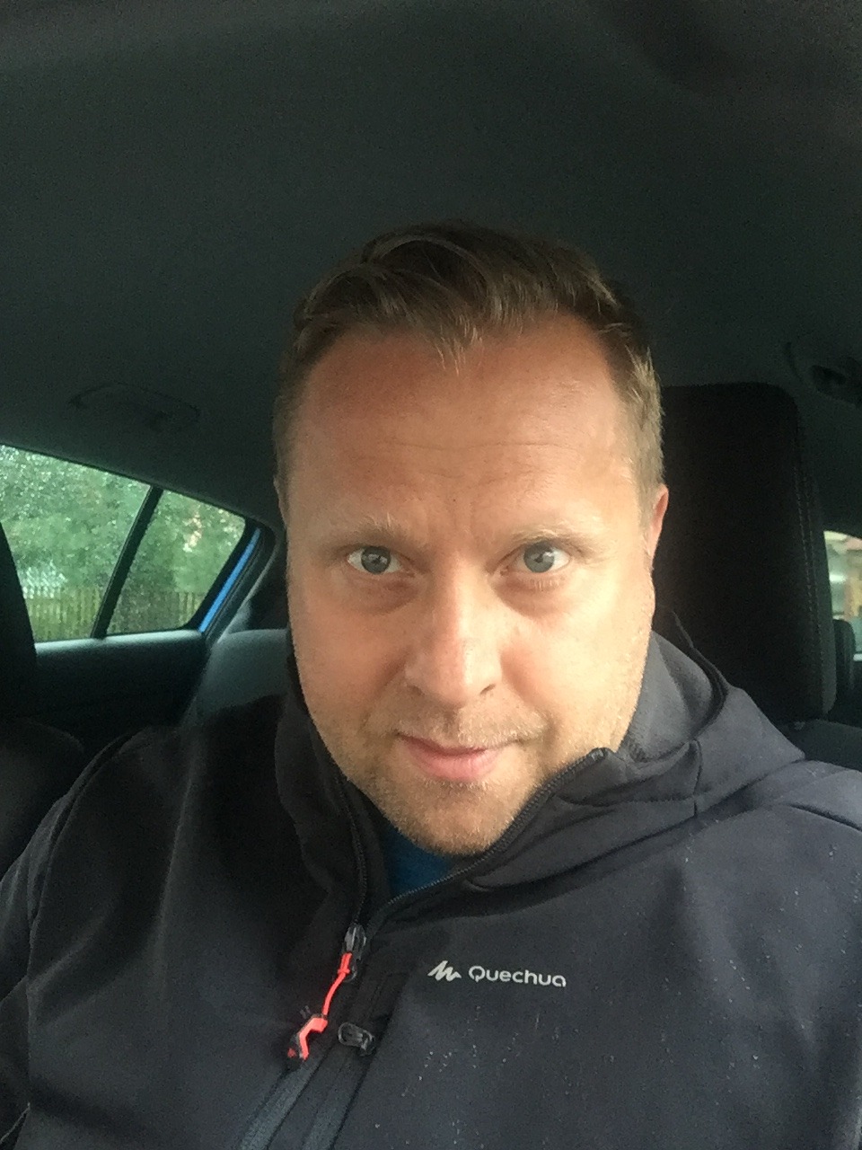 Mężczyzna w aucie, ubrany w czarną kurtkę Quechua, patrzy w obiektyw. Widoczne drzewa za oknem.