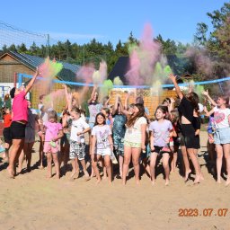 Invictus Przemysław Gałaszewski - Grupka roześmianych dzieci na piaszczystym boisku radośnie wyrzuca w górę kolorowe proszki podczas zabawy w słoneczny dzień. W tle siatka do siatkówki i budynek.