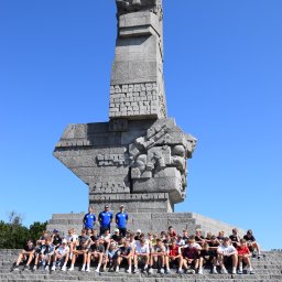 Invictus Przemysław Gałaszewski - Grupa młodzieży z dwoma trenerami pozuje na schodach przed Pomnikiem Obrońców Wybrzeża w Gdańsku. Błękitne niebo w tle.