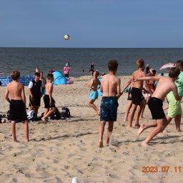 Invictus Przemysław Gałaszewski - Grupa dzieci gra w siatkówkę na piaszczystej plaży nad morzem, w tle widoczne parawany i parasole, a w górze unosi się piłka do siatkówki.