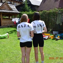 Invictus Przemysław Gałaszewski - Dwie dziewczyny w białych koszulkach z imionami 'MAJA' i 'WIKA' na plecach, stoją tyłem na trawniku w słoneczny dzień. W tle drewniana altana i zabawki.