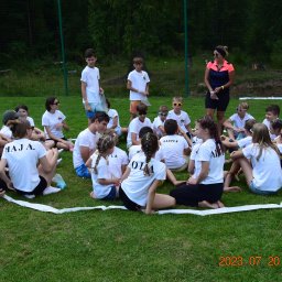 Invictus Przemysław Gałaszewski - Grupa dzieci i opiekunka siedzą na trawie w kręgu wyznaczonym taśmą. Uczestnicy w białych koszulkach z imionami. Zdjęcie z kolonii lub obozu.