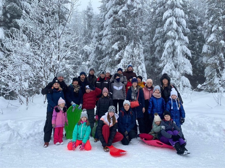 Grupa ludzi z sankami w zimowym lesie, prawdopodobnie uczestnicy wycieczki lub obozu zimowego. Śnieżne drzewa w tle, pochmurne niebo.