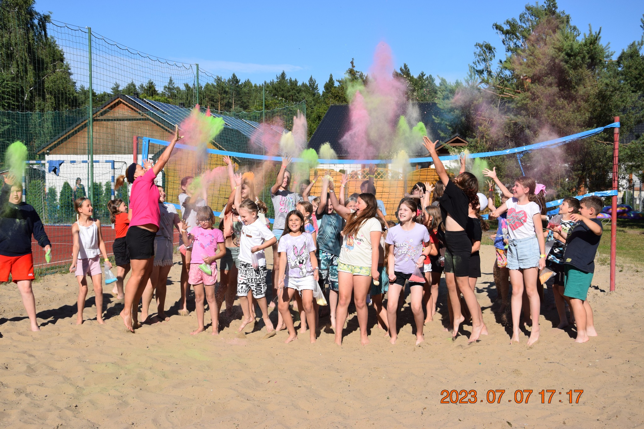 Grupka roześmianych dzieci na piaszczystym boisku radośnie wyrzuca w górę kolorowe proszki podczas zabawy w słoneczny dzień. W tle siatka do siatkówki i budynek.