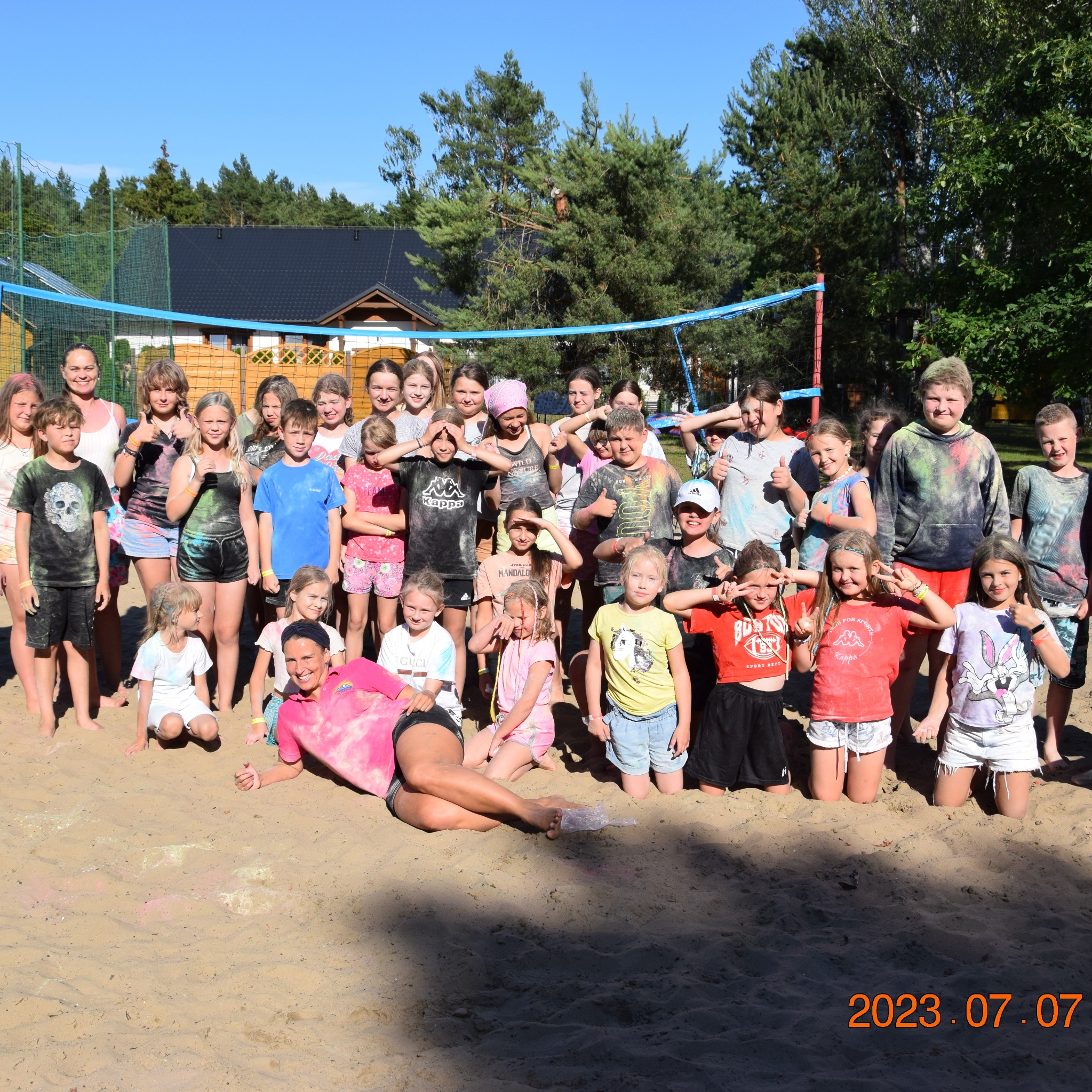 Grupa dzieci i opiekunka po zabawie kolorami na piaszczystym boisku z siatką do siatkówki w tle, w słoneczny dzień. Data 2023.07.07 w prawym dolnym rogu.