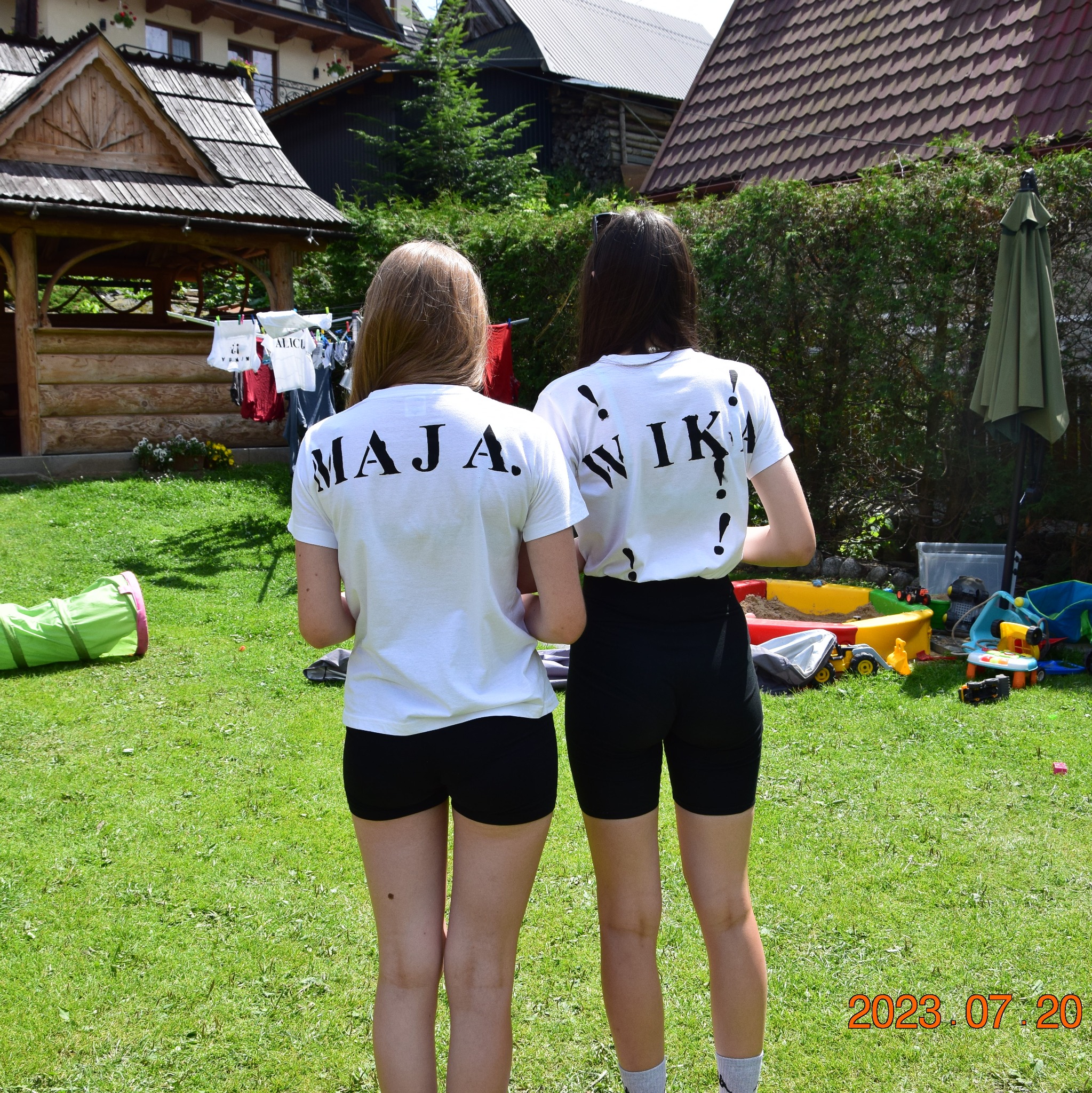 Dwie dziewczyny w białych koszulkach z imionami 'MAJA' i 'WIKA' na plecach, stoją tyłem na trawniku w słoneczny dzień. W tle drewniana altana i zabawki.