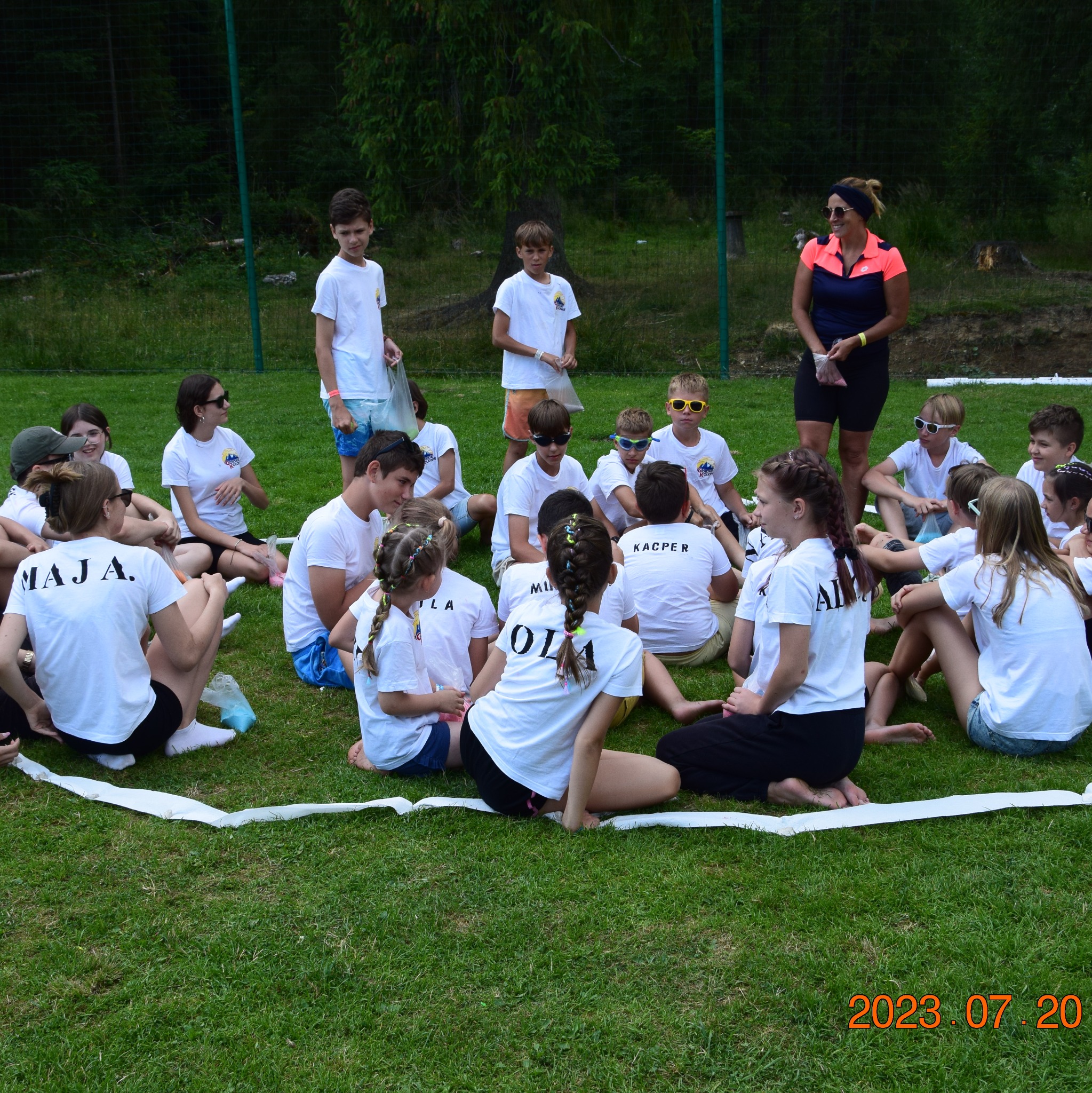 Grupa dzieci i opiekunka siedzą na trawie w kręgu wyznaczonym taśmą. Uczestnicy w białych koszulkach z imionami. Zdjęcie z kolonii lub obozu.