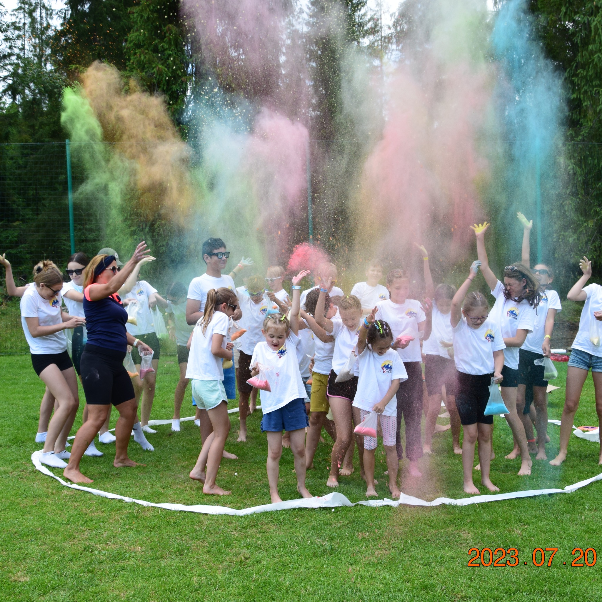Grupa dzieci i dorosłych w białych koszulkach bawi się proszkami Holi na zielonej trawie, tworząc kolorową chmurę. Widoczna data 2023.07.20.