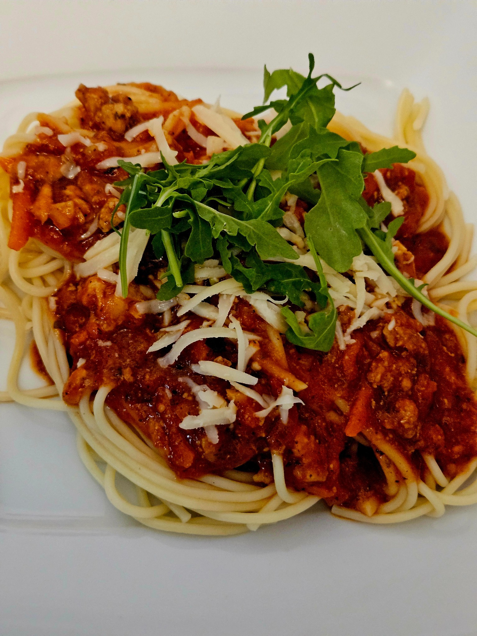 Apetyczne spaghetti z sosem bolońskim, posypane serem i świeżą rukolą, podane na białym talerzu. Gotowe danie.