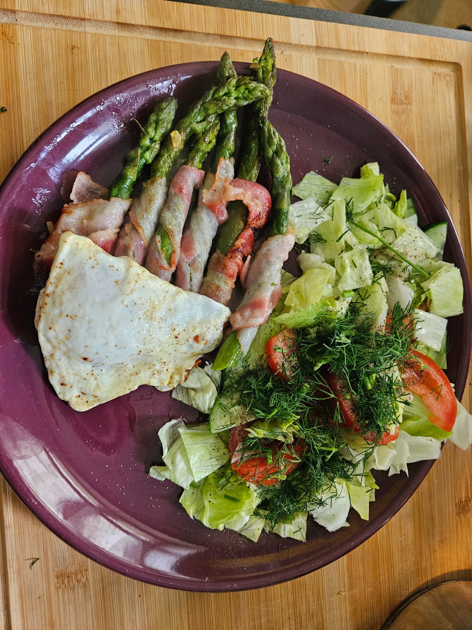 Fioletowy talerz z grillowanymi szparagami w boczku, jajkiem sadzonym i sałatką z pomidorami i koperkiem na drewnianej desce. Danie gotowe do spożycia.