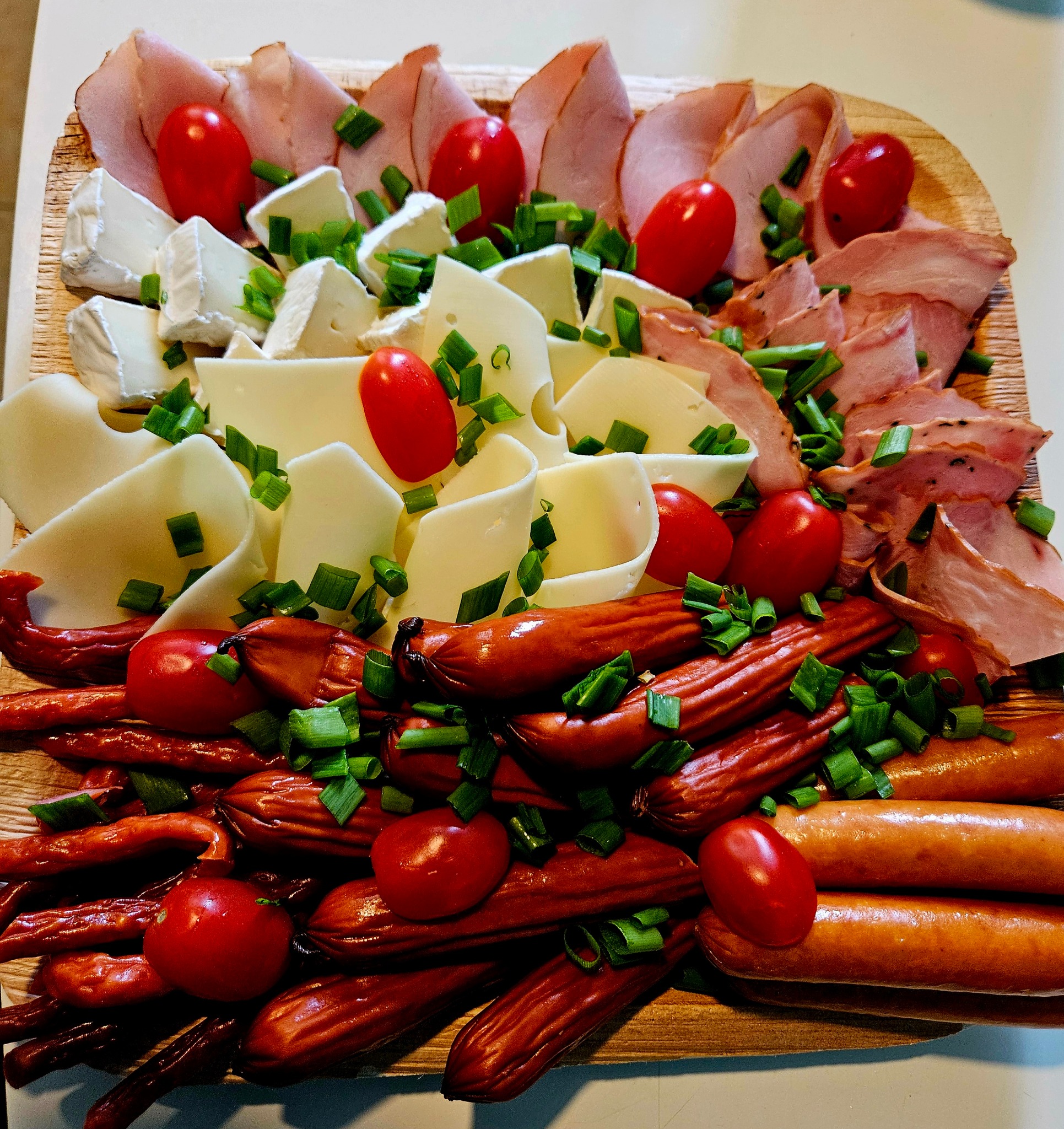 Drewniana deska z wykwintną kompozycją serów, wędlin, kiełbasek i pomidorków cherry, posypana szczypiorkiem. Idealna przekąska na przyjęcie.
