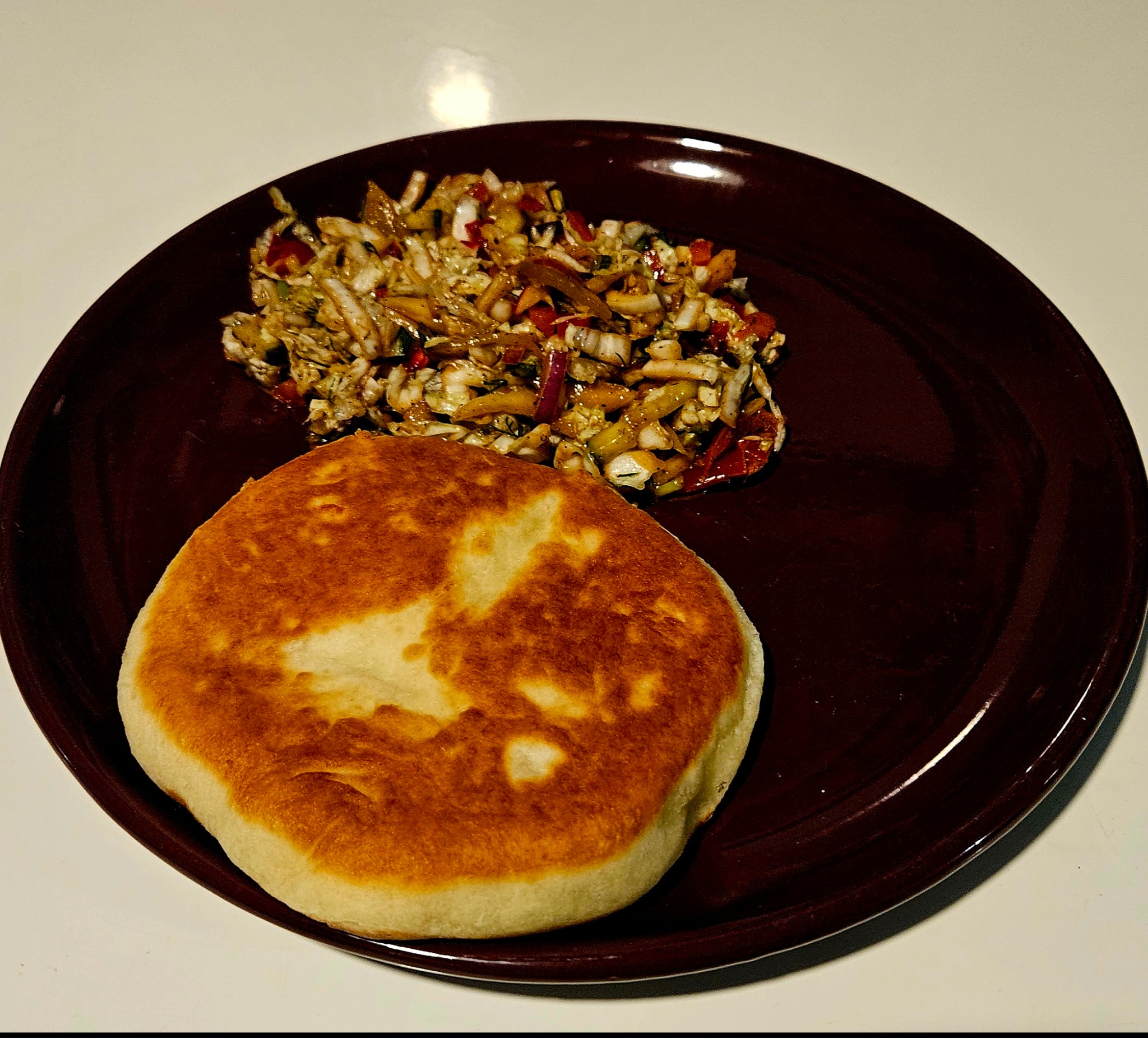 Okrągły placek obok sałatki warzywnej na brązowym talerzu, oświetlony punktowo, na jasnym tle. Potrawa gotowa do spożycia, prezentacja dań cateringowych.