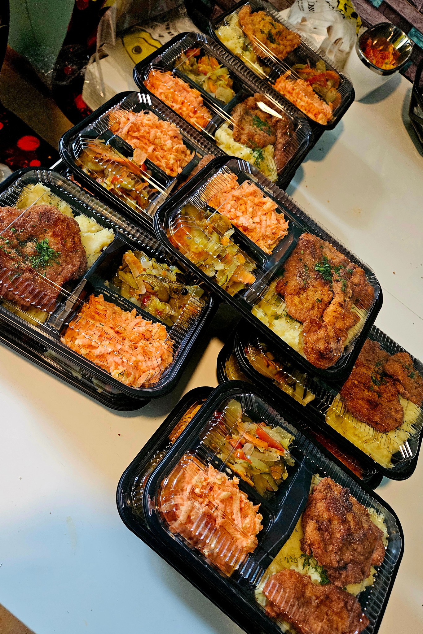 Zestaw obiadowy w czarnych pojemnikach z przezroczystym wieczkiem: kotlet, puree, surówka z marchewki i warzywa duszone. Gotowe porcje cateringu.