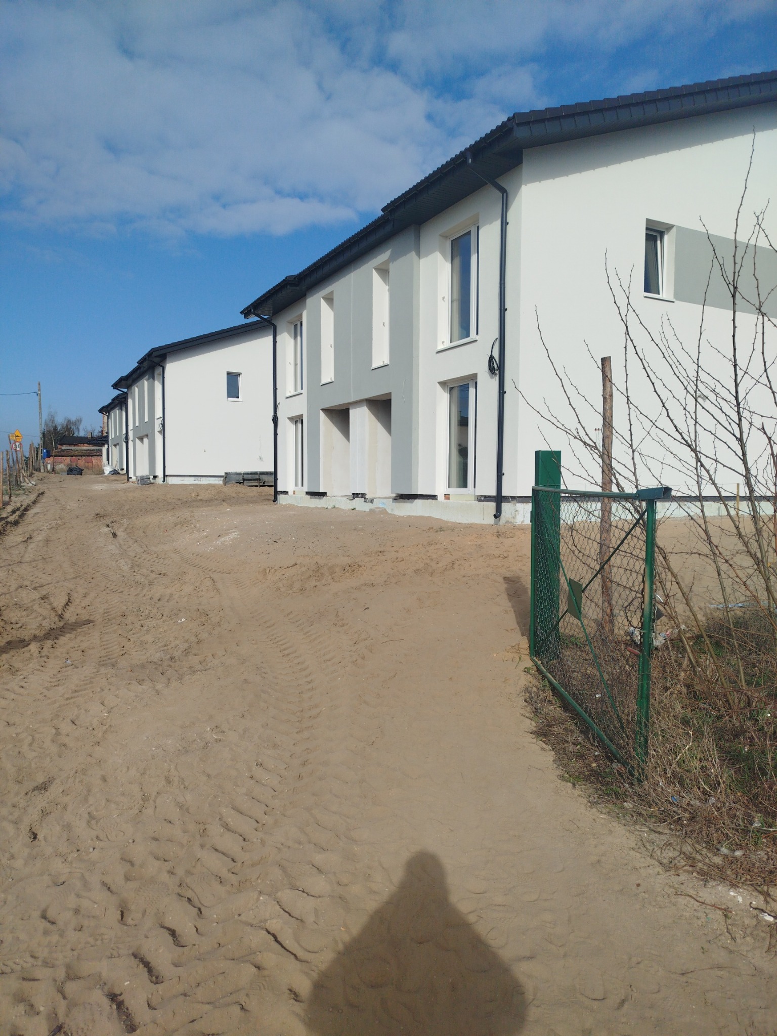 Nowoczesne szeregówki w jasnej kolorystyce, z czarnymi akcentami dachu i rynien, usytuowane w surowym otoczeniu piaszczystego placu budowy i zielonego ogrodzenia.
