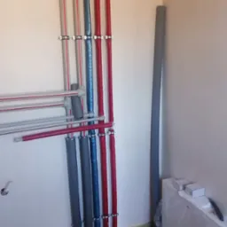 Pionowe rury instalacyjne w kolorach czerwonym, niebieskim i szarym, przymocowane klamrami do ściany. Dodatkowe poziome rury z szarymi złączkami.