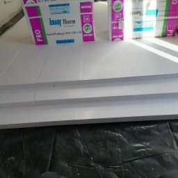 Jacek ŁUCZAK - Układanie białych płyt styropianowych Knauf Therm na czarnej folii, przygotowanie pod ogrzewanie podłogowe. Widoczne paczki produktu.