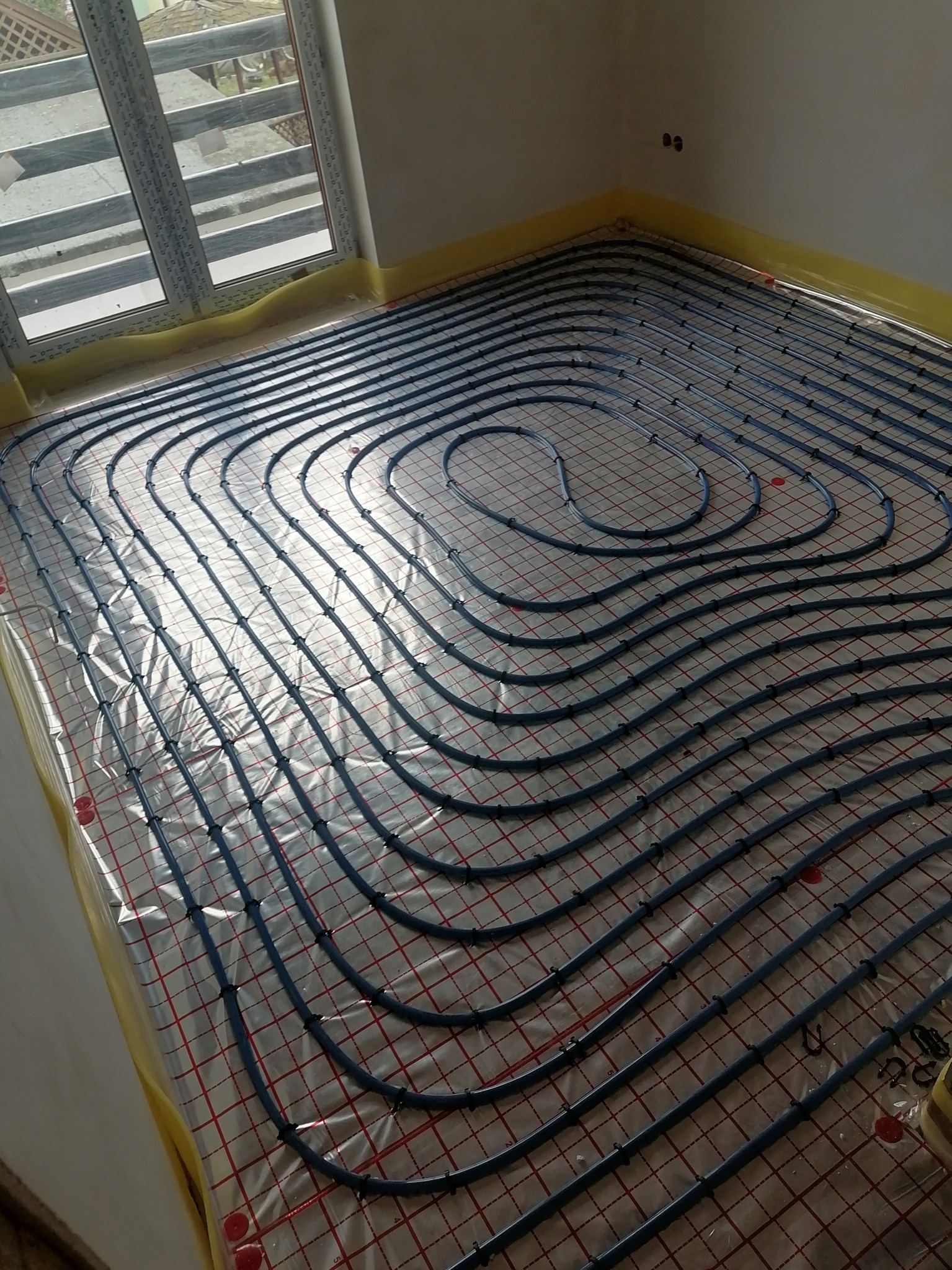 Niebieskie rury ogrzewania podłogowego ułożone spiralnie na folii z czerwoną siatką, widok z góry, przy oknie. Żółta folia ochronna na ścianach.