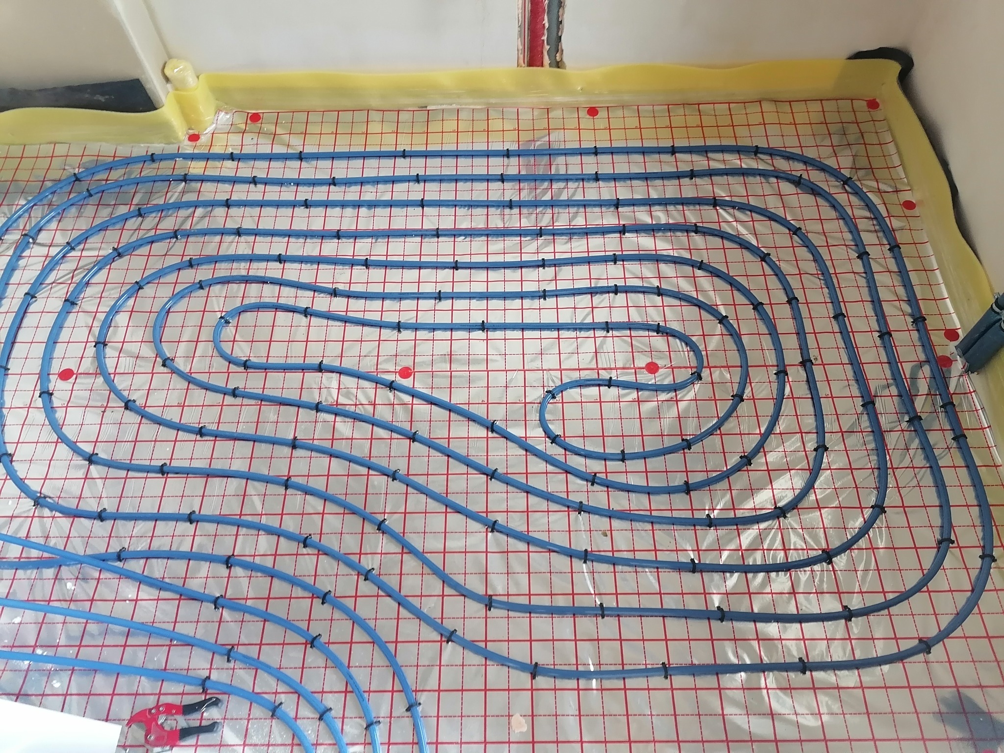Instalacja ogrzewania podłogowego: niebieskie rurki ułożone spiralnie na folii z czerwoną siatką, przymocowane czarnymi uchwytami. Widoczne narzędzie do cięcia rur.