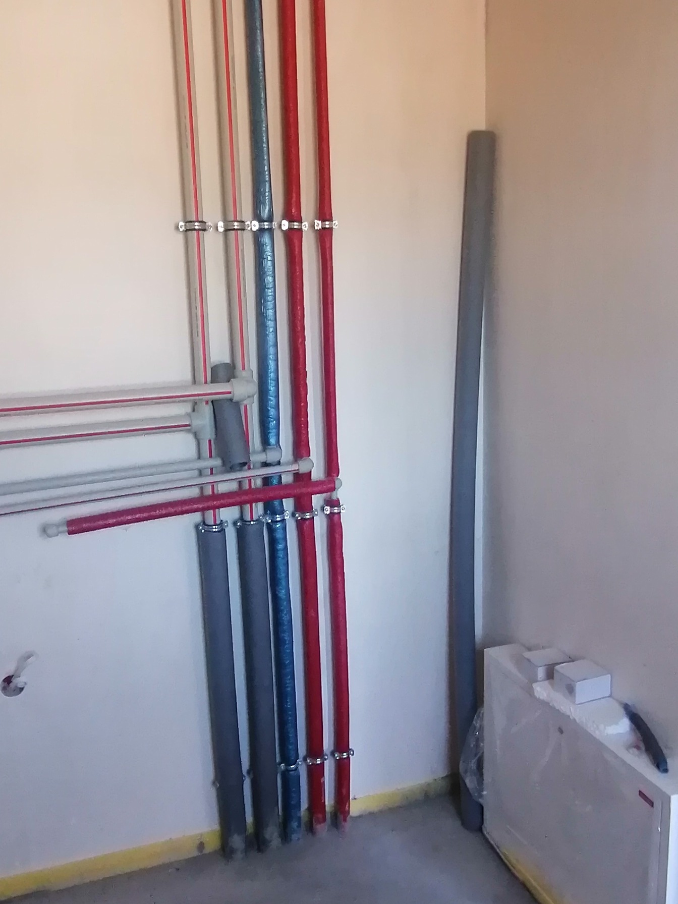 Pionowe rury instalacyjne w kolorach czerwonym, niebieskim i szarym, przymocowane klamrami do ściany. Dodatkowe poziome rury z szarymi złączkami.