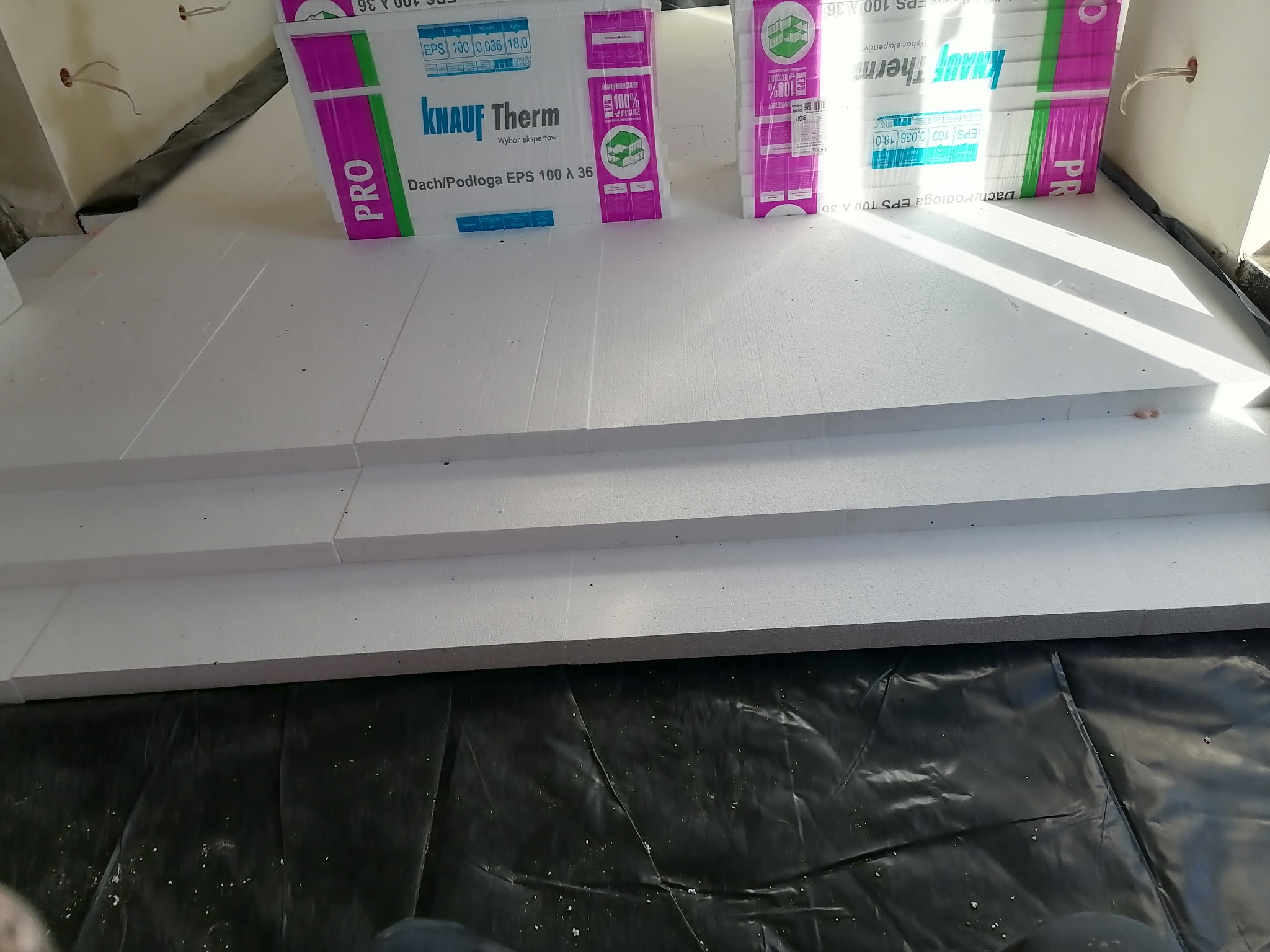 Układanie białych płyt styropianowych Knauf Therm na czarnej folii, przygotowanie pod ogrzewanie podłogowe. Widoczne paczki produktu.