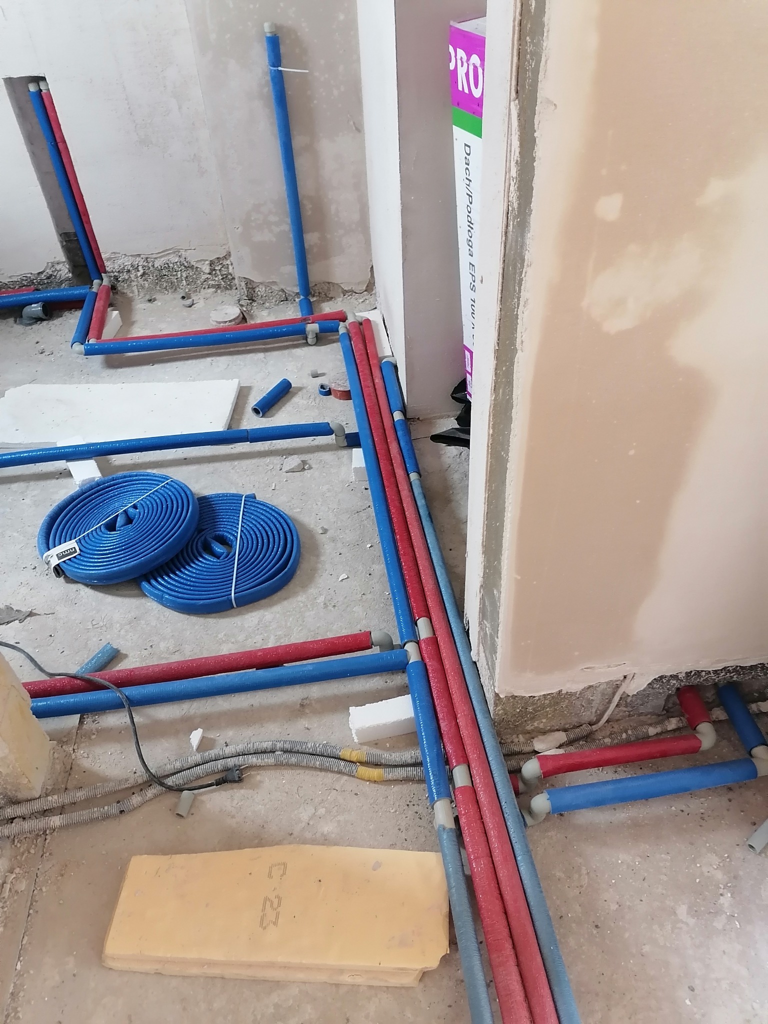 Instalacja wodna w trakcie montażu: niebieskie i czerwone rury prowadzone wzdłuż ścian, zwoje rur na podłodze, widoczne elementy izolacji. Surowy stan wykończenia.