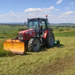 Czerwony traktor Massey Ferguson 5445 z żółtym pługiem na polu po koszeniu trawy, w tle zielony krajobraz i błękitne niebo z chmurami.