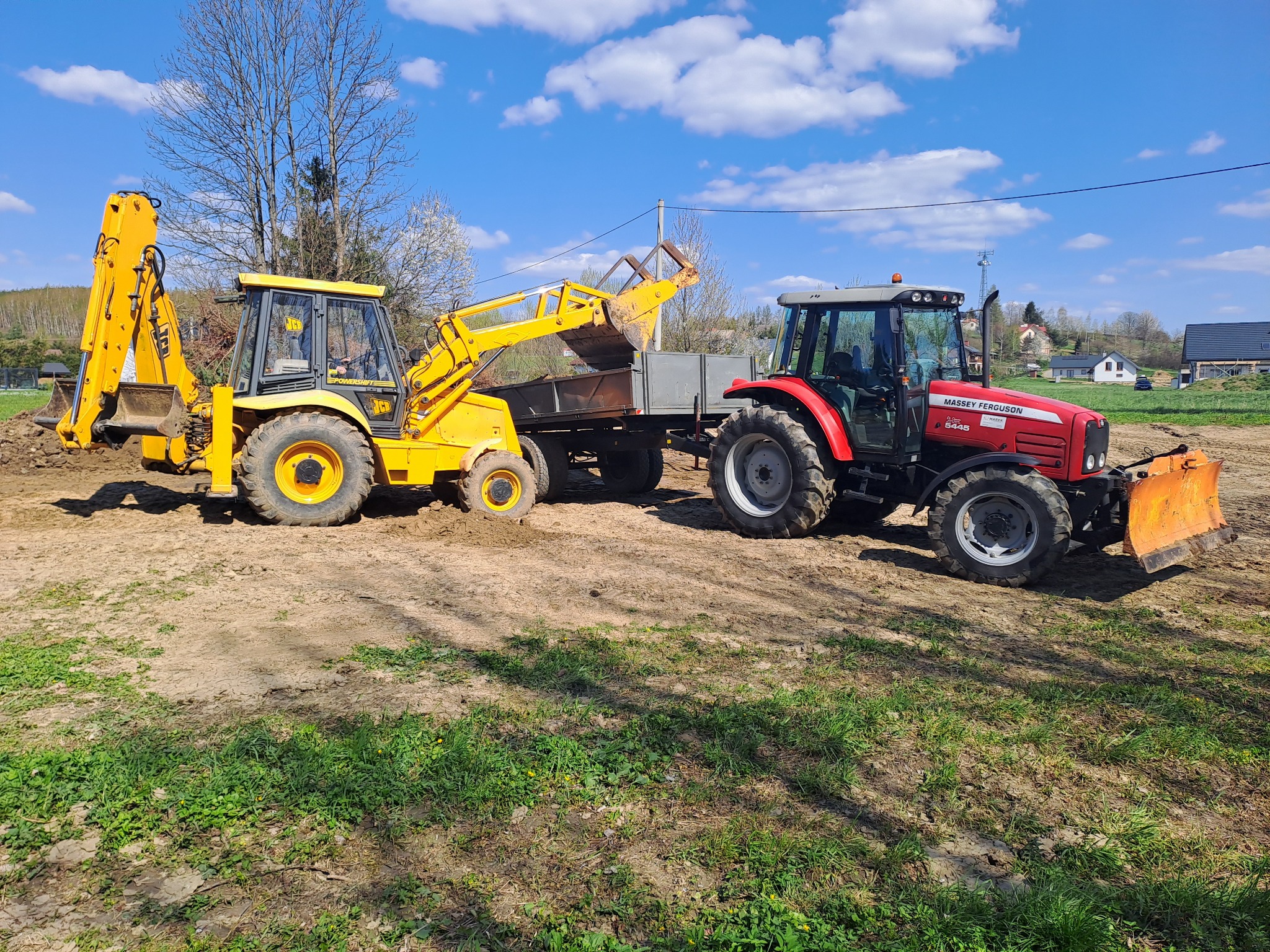 Żółto-czarna koparko-ładowarka JCB ładuje ziemię do przyczepy ciągniętej przez czerwony traktor Massey Ferguson na placu budowy w słoneczny dzień.