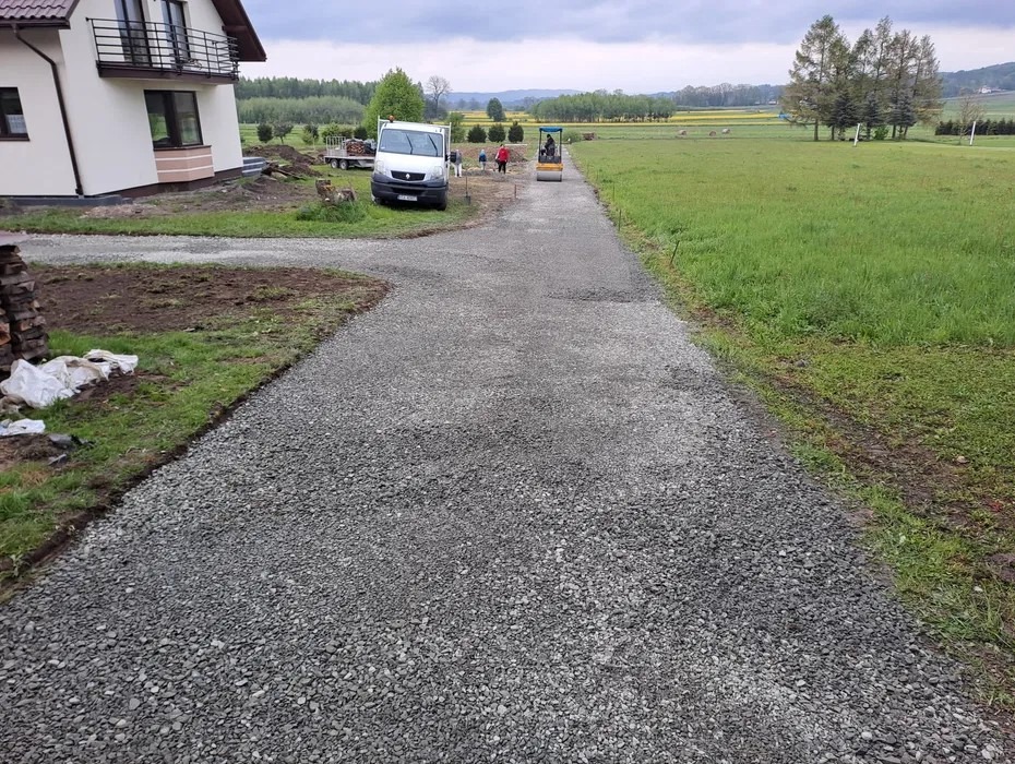 Utwardzona droga dojazdowa z szarego kruszywa prowadząca do domu, w tle widać walec drogowy i samochód dostawczy. Krajobraz wiejski.