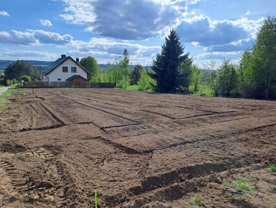 Przygotowany teren pod budowę z wyznaczonymi fundamentami, widok na dom i zielone otoczenie w słoneczny dzień.