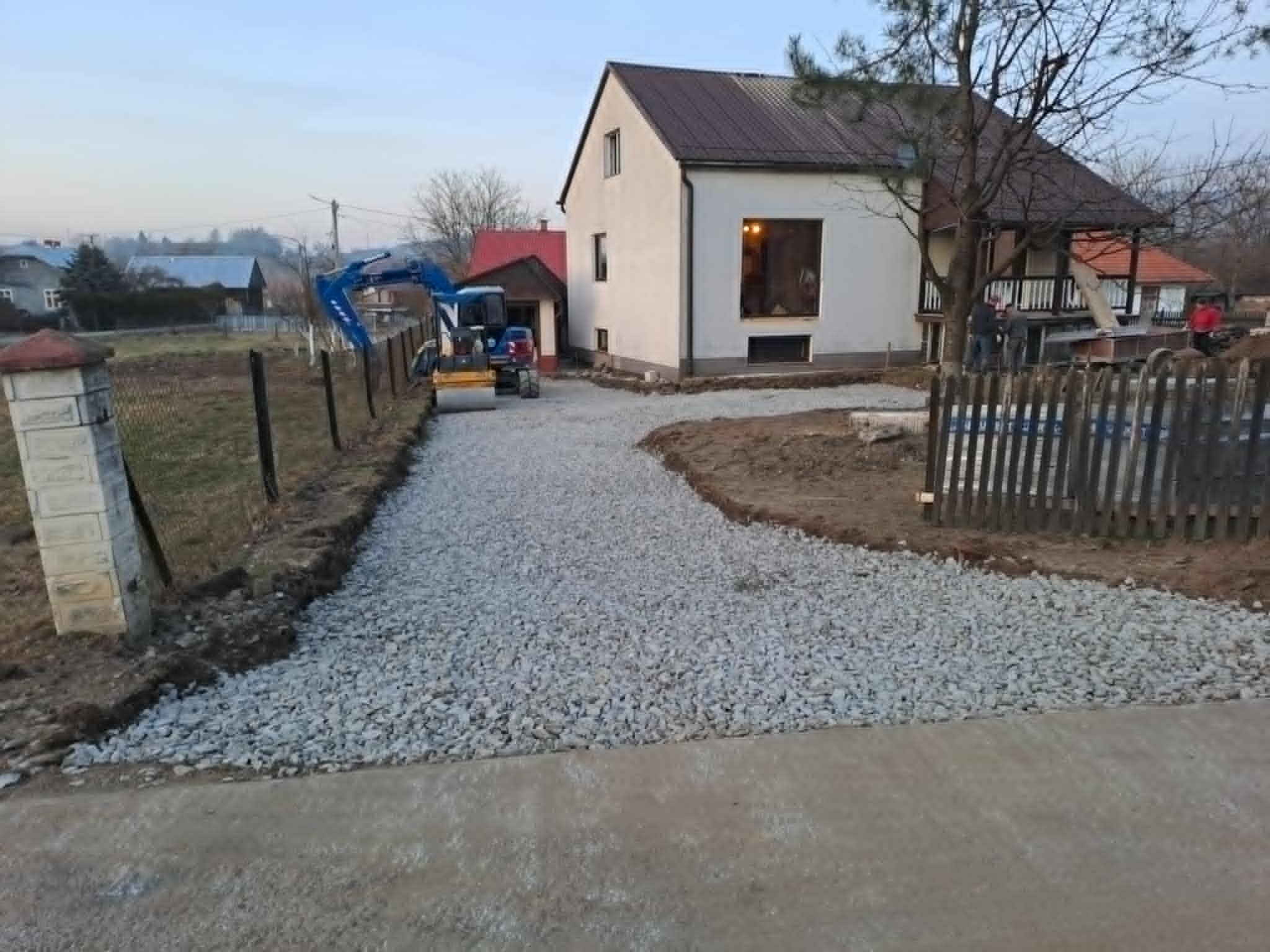 Utwardzona żwirem droga do domu z użyciem walca drogowego. W tle dom z brązowym dachem i grupa osób przy drewnianym ogrodzeniu.