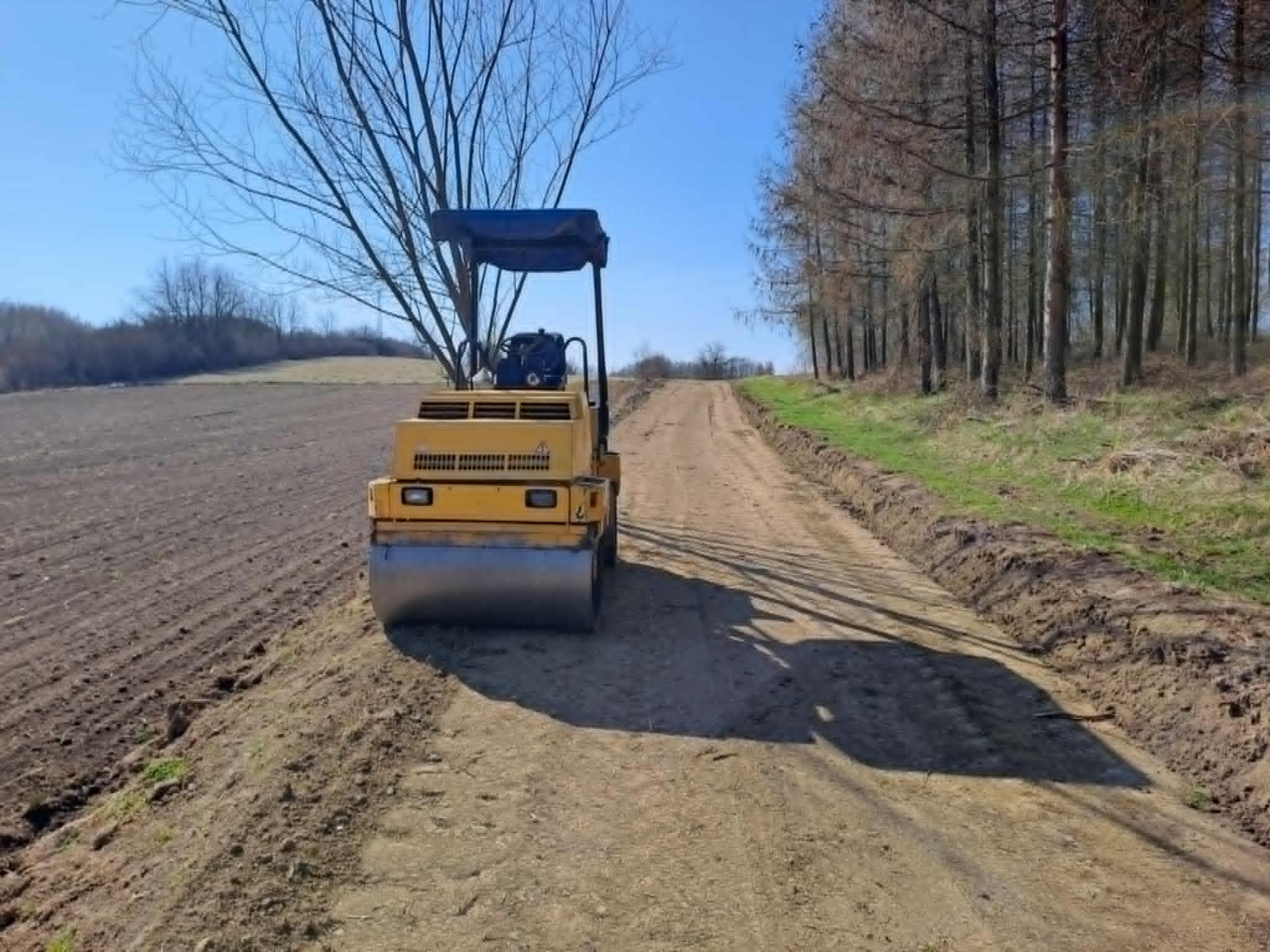 Żółty walec drogowy ubija gruntową drogę na tle pola i lasu w słoneczny dzień. Widoczne ślady opon i cień maszyny na ubitej nawierzchni.