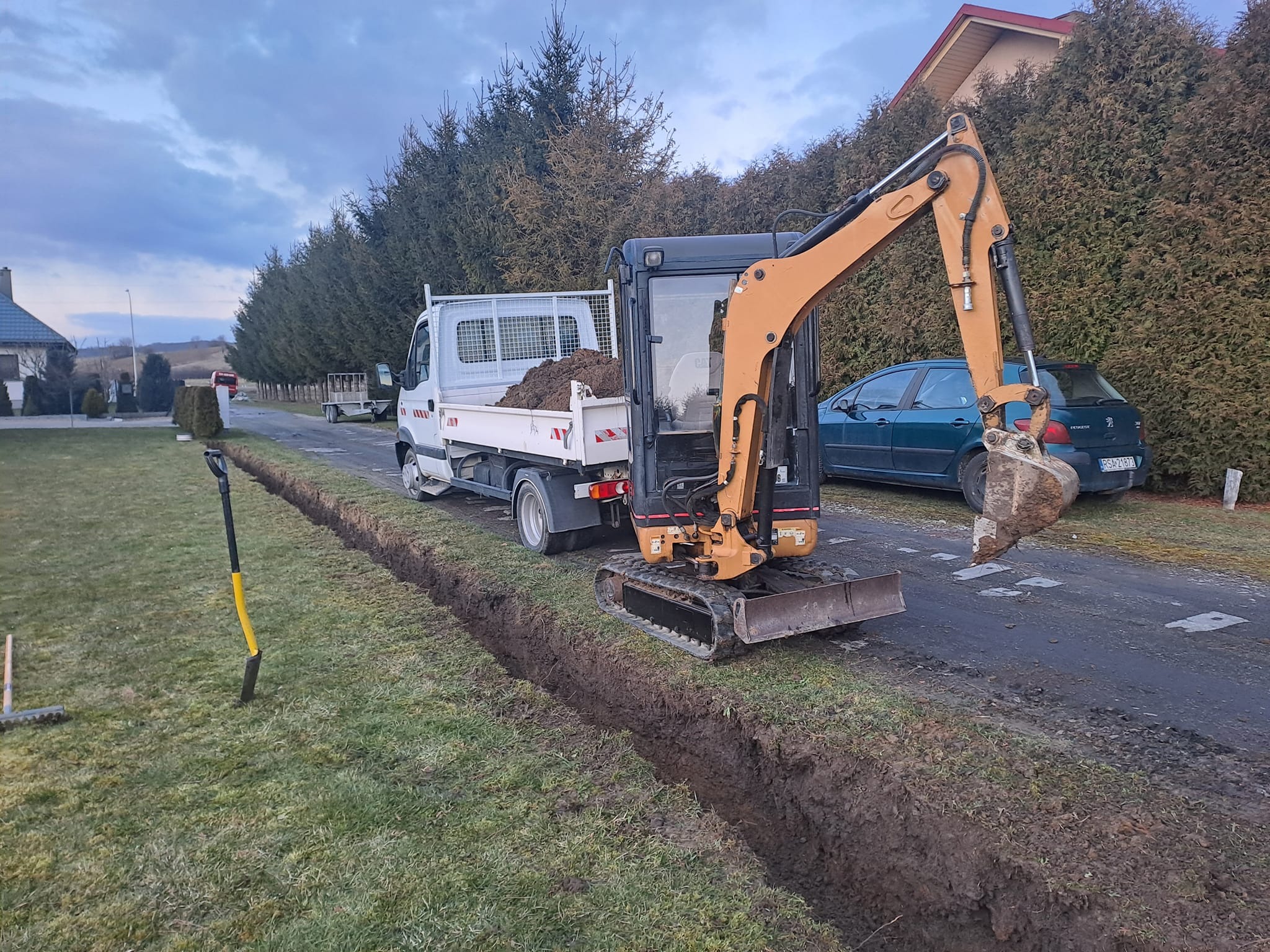 Mini koparka kopie wąski rów obok drogi, wykopana ziemia leży na pace białej ciężarówki. W tle zielony samochód i tuje.