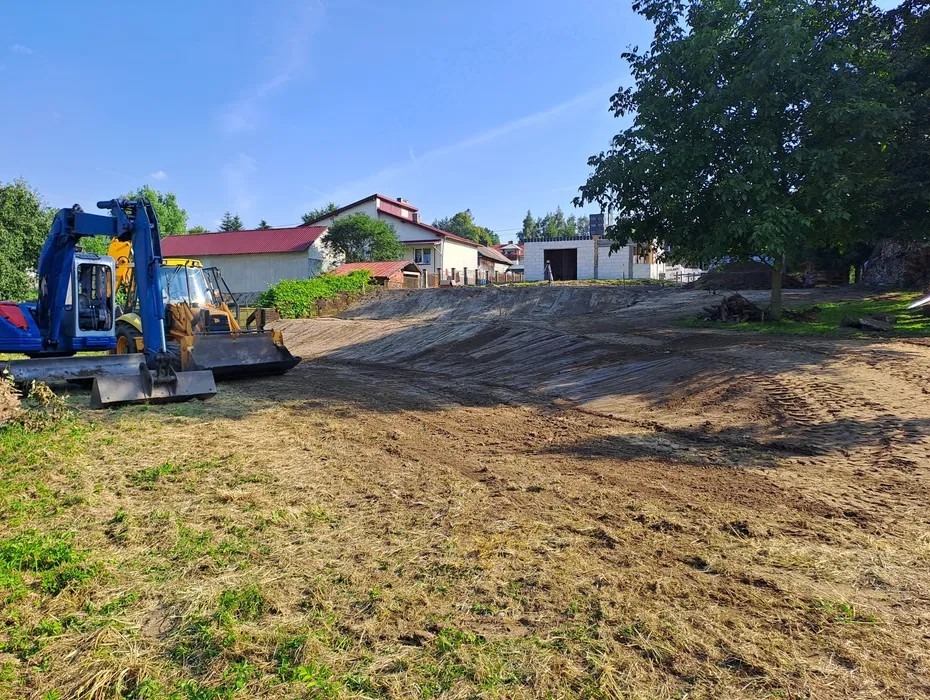 Niwelacja terenu pod budowę: wyrównany grunt, widoczne maszyny budowlane (koparki) i budynek w tle. Słoneczny dzień, teren przygotowany pod inwestycję.