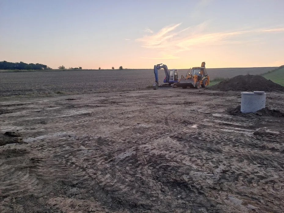 Nierówny teren po robotach ziemnych z widocznym wykopem pod szambo, obok koparka i koparko-ładowarka. W tle zachodzące słońce.
