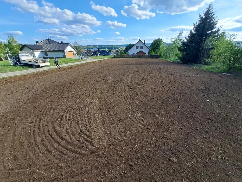 Przygotowany, wyrównany teren pod budowę lub ogród na tle domów i błękitnego nieba. Widoczne ślady po maszynach rolniczych.
